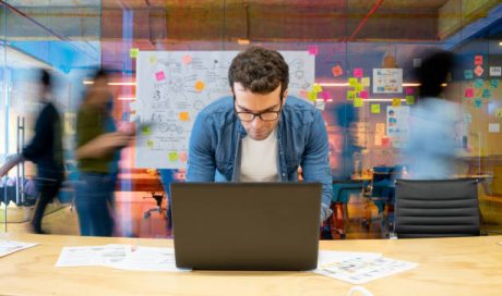 istockphoto-1332176260-612×612 Latin American man working at a creative office using his computer and people moving at the background - place of work concepts